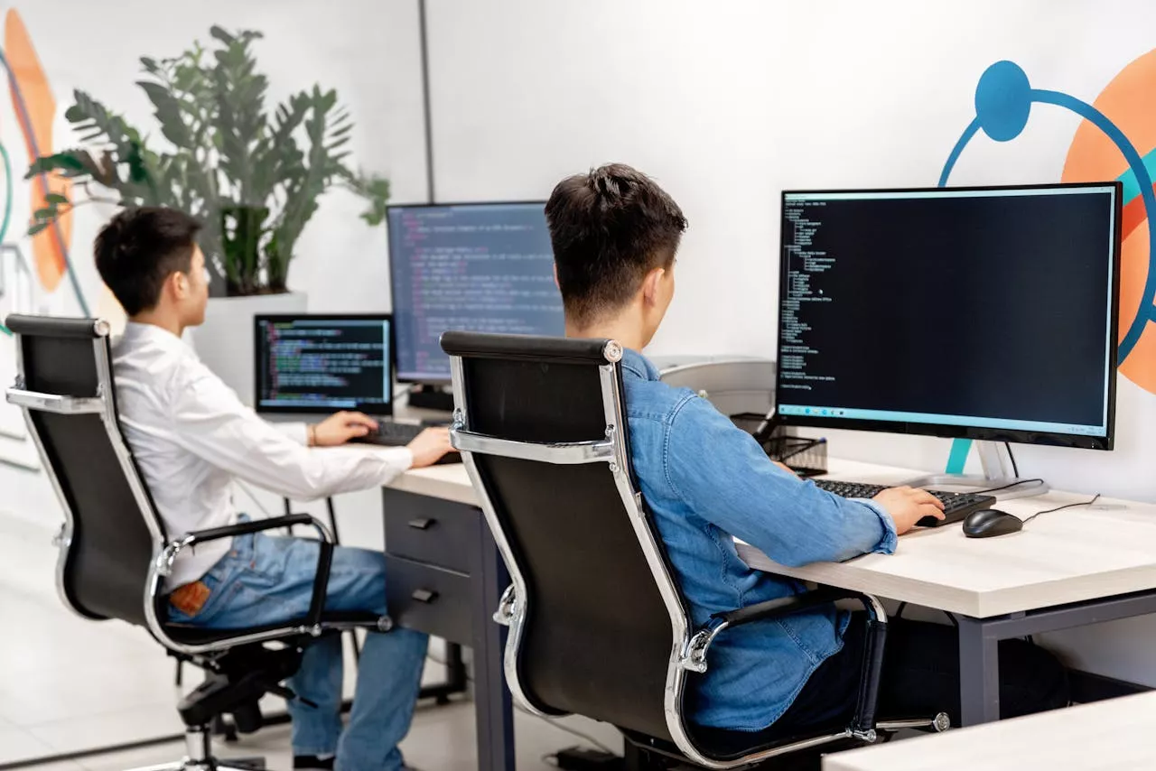 journey-01 Two male developers at desks programming in a modern office workspace with large monitors.